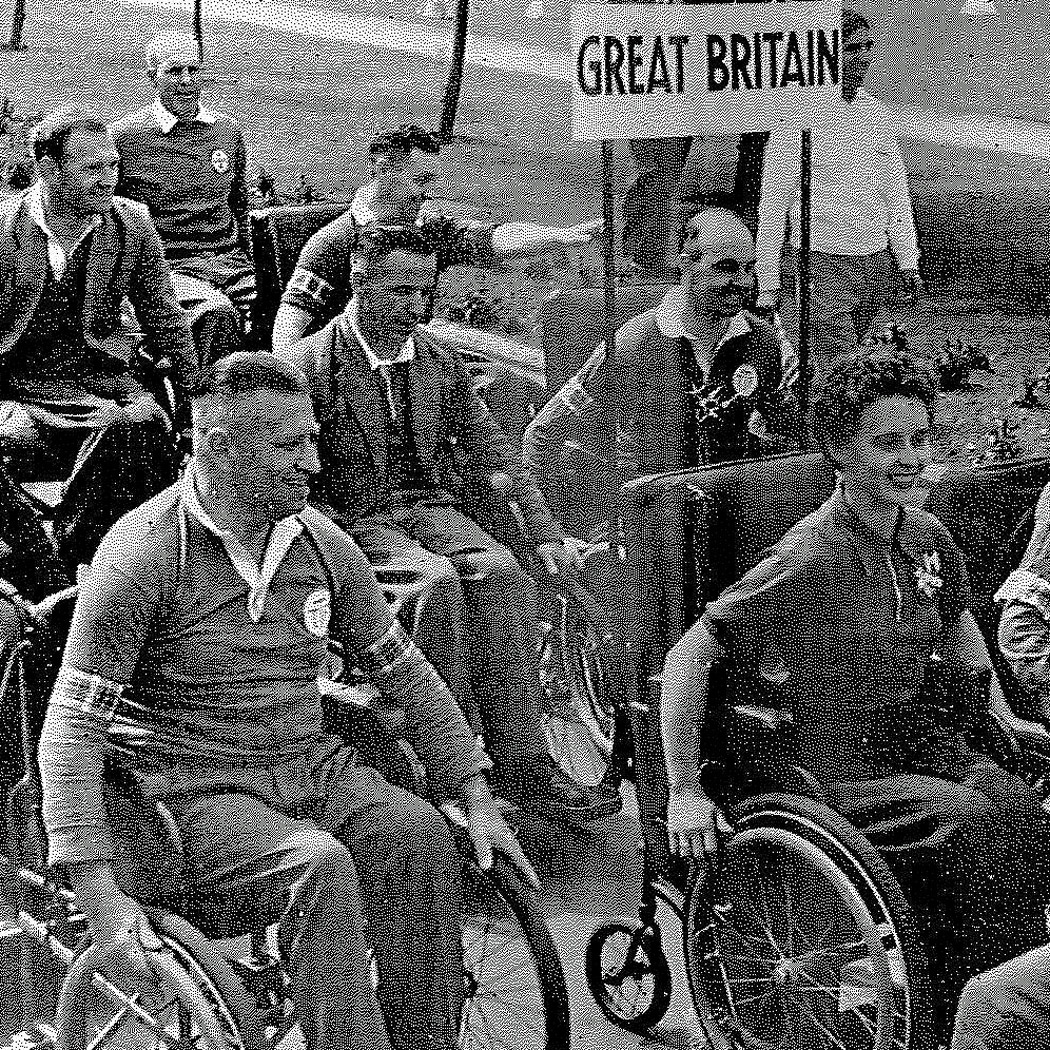 Grainy black and white photo of wheelchair athletes in a parade with a banner saying Great Britain. 