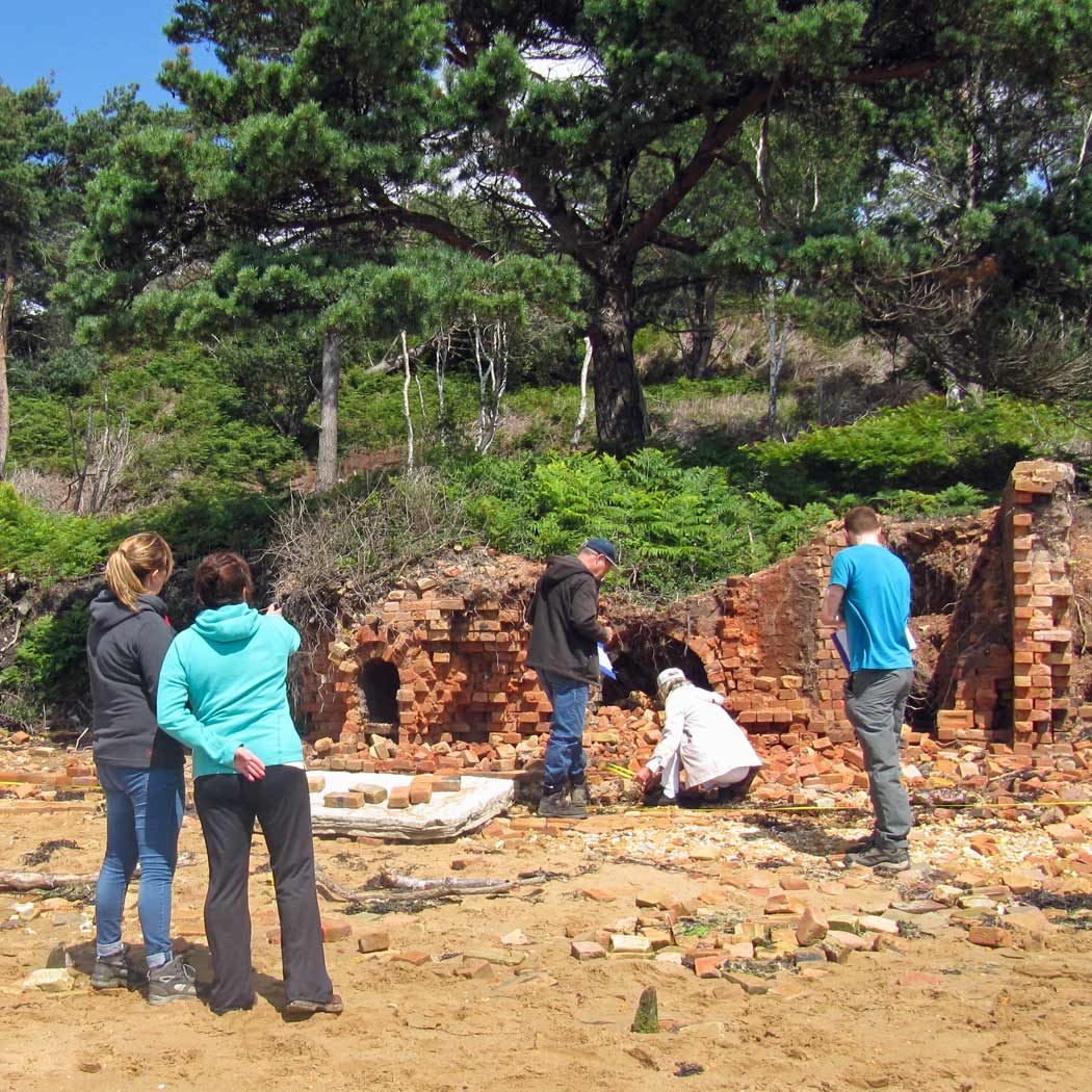 People standing on a beach looking at a collapsing brick wall, part of a kiln.