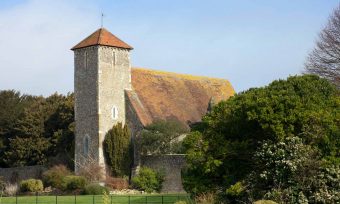 Old stone church with square spire, red tiled roof and bushes in the foreground.