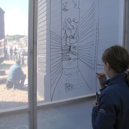 Young girl drawing a picture on a transparent material, with beach huts outside.