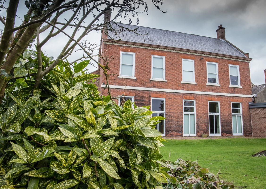 Red brick building behind some grass and a bush. 