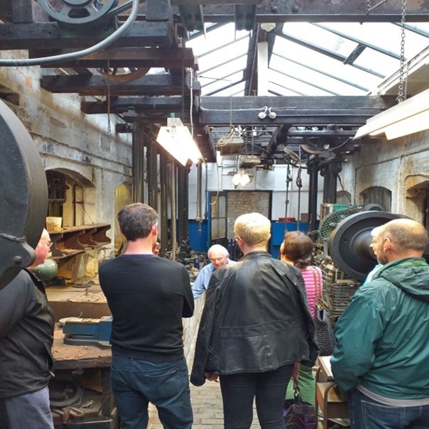 A group of people stand in an old industrial workshop, watching a man showing them something.