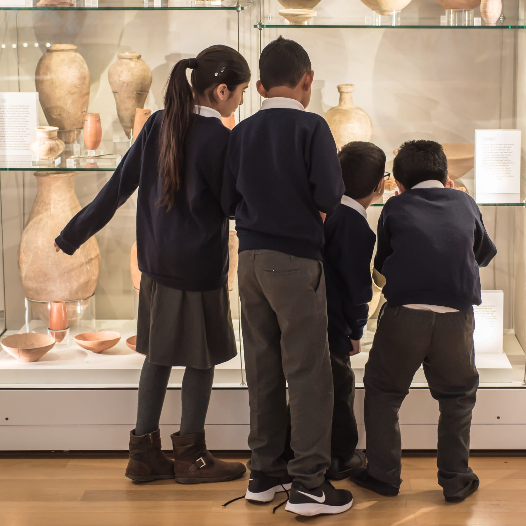 Four school children in uniform look into a museum display case containing Egyptian ceramics