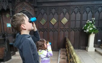 Two children stand in front of wooden carved panels, using binoculars to look at something in the distance.